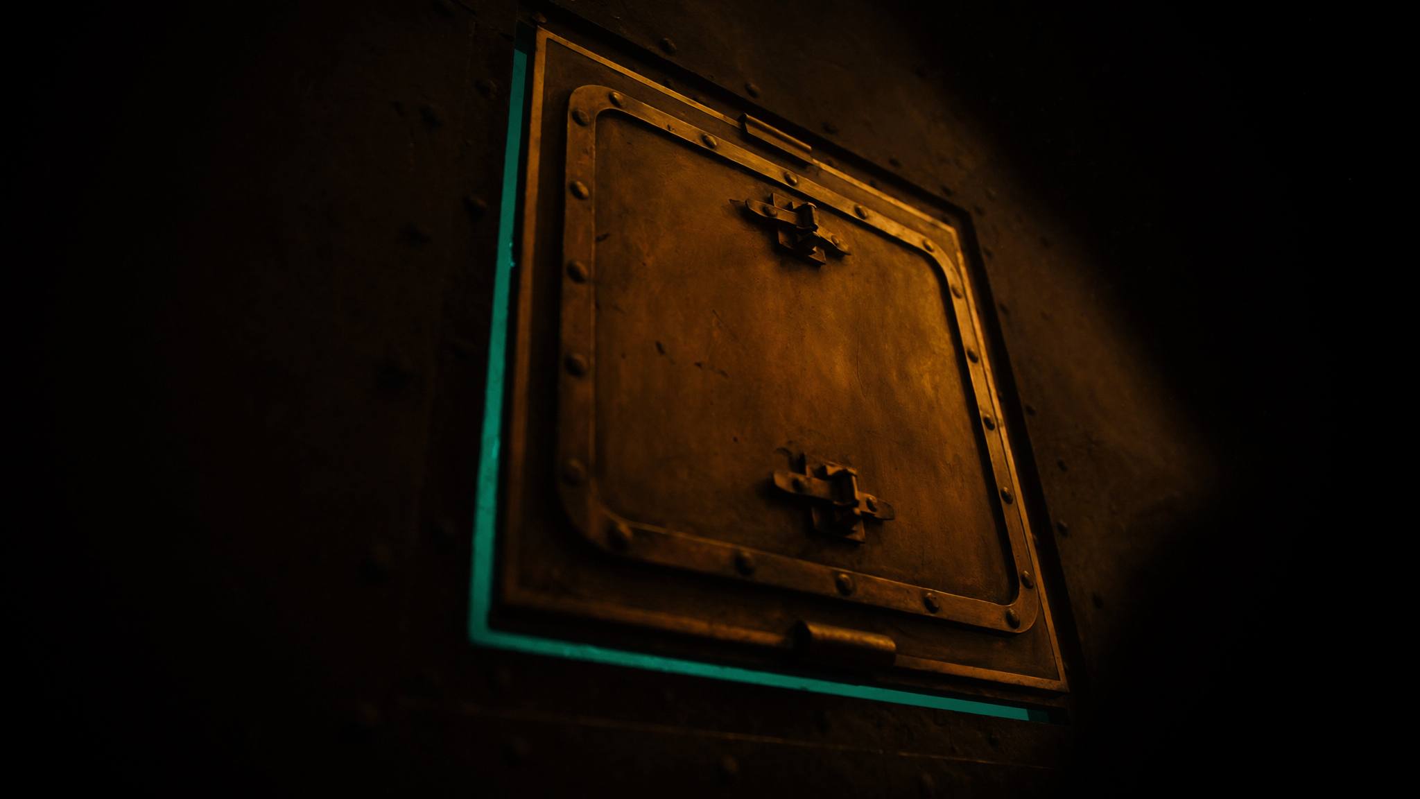 Schwere Messing-Schottentür in dunkler Wand, teal leuchtender Spalt am Rahmen